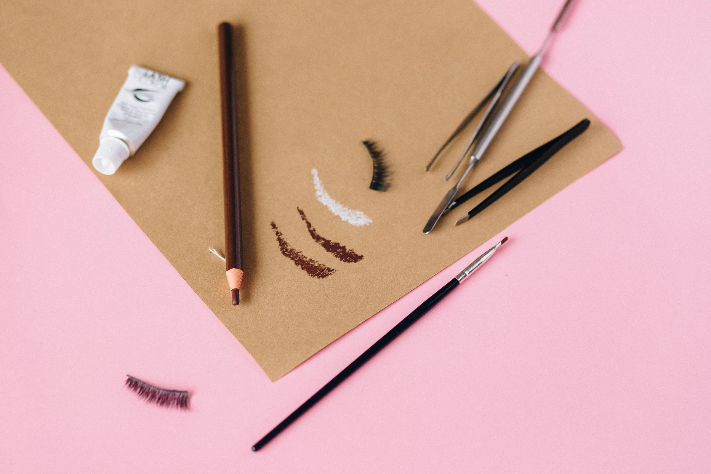 Brow pencils, tweezers and false lashes laid out on pink and kraft paper